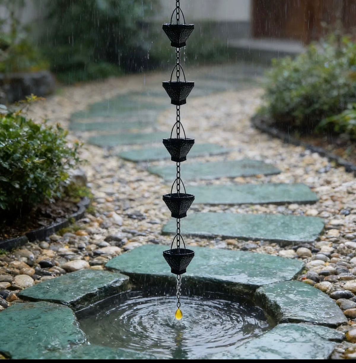 Black hammered cup rain chain guiding rainwater into a stone basin on a garden path lined with stepping stones and pebbles