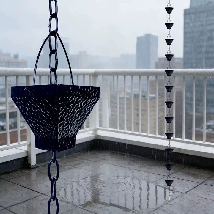 Black hammered cup rain chain hanging from a balcony railing during heavy rain, with a city skyline in the background